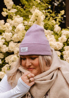 Une personne portant un bonnet douillet Mimi & August Lilac Beanie et une écharpe beige se tient devant des buissons fleuris blancs.