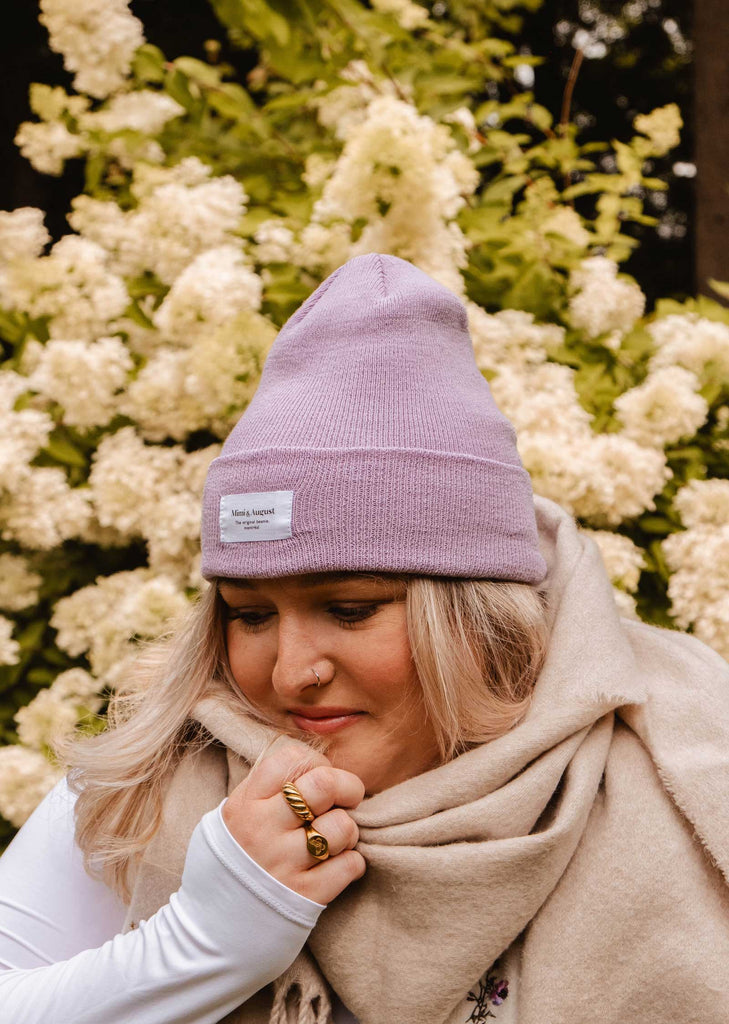 A person wearing a cozy Mimi & August Lilac Beanie and a beige scarf stands in front of white flowering bushes.