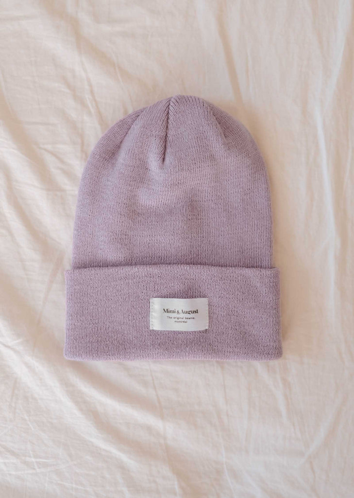 Le bonnet Lilac de Mimi & August, un tricot lilas avec un bord plié et une étiquette blanche rectangulaire sur un fond de tissu beige, est l'accessoire douillet parfait. Ce tricot unisexe est à la fois élégant et polyvalent.