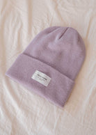 Le bonnet lilas de Mimi & August est doté d'une étiquette rectangulaire blanche, joliment présentée sur un fond de tissu beige doux, ce qui en fait un accessoire idéal pour tout le monde.