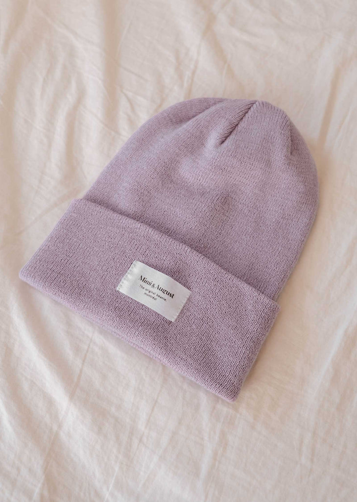 A cozy lilac beanie from Mimi & August featuring a white rectangular label, beautifully displayed on a soft beige fabric background, making it an ideal accessory for everyone.