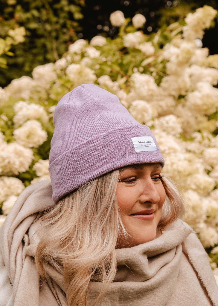 A person wearing a Lilac Beanie from Mimi & August and a cream unisex knit scarf is standing in front of white flowering shrubs.