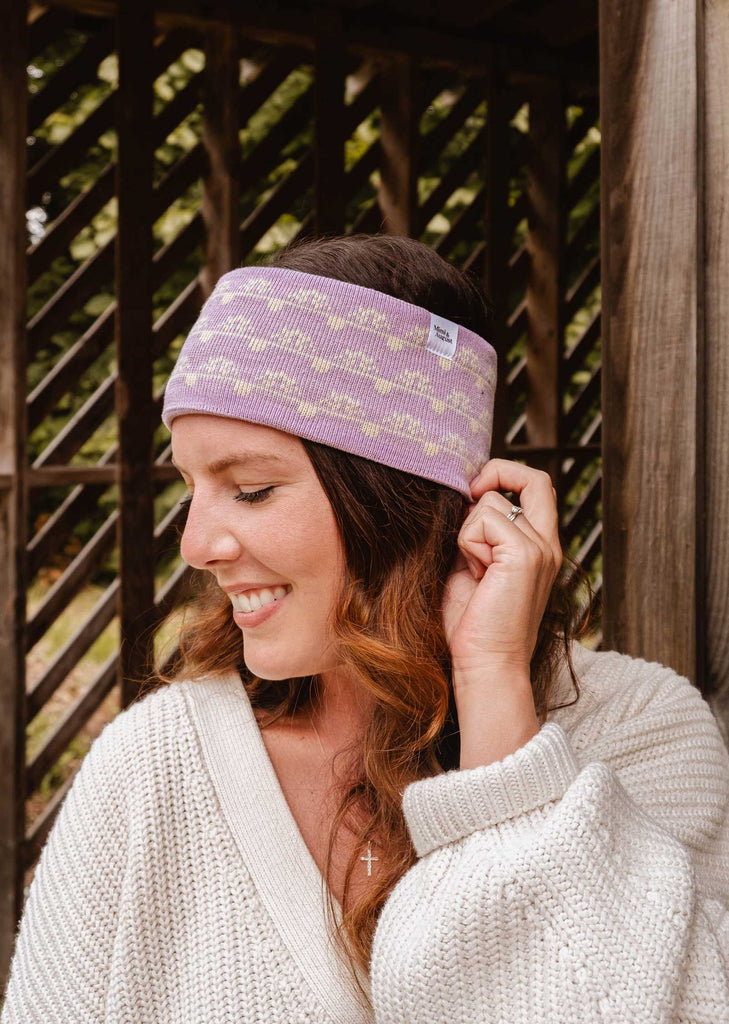 A woman with long hair, smiling and exuding cozy warmth, stands in front of a wooden structure wearing a white sweater and the Lilac Bliss Headband from Mimi & August. She is radiating cold-weather elegance as she holds the headband with her left hand.