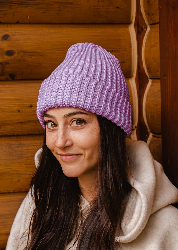 A woman wearing a Lilac Chunky Beanie in front of a log cabin.