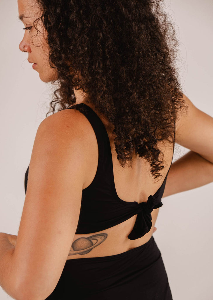 A woman with curly hair and a visible nose piercing is wearing a Mimi & August Lima Black Bralette Bikini Top with an adjustable back tied in a bow, exposing a tattoo of Saturn on her left side. She is looking to the left.