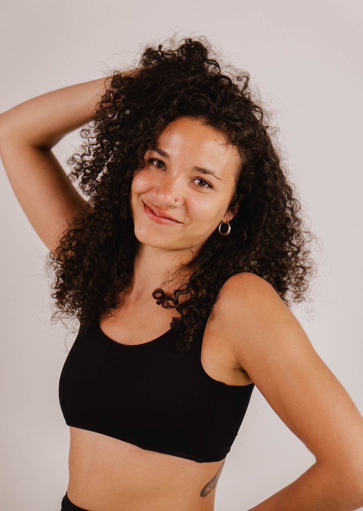 A woman with curly hair wearing a Mimi & August Lima Black Bralette Bikini Top with removable pads poses with one arm behind her head, smiling at the camera.