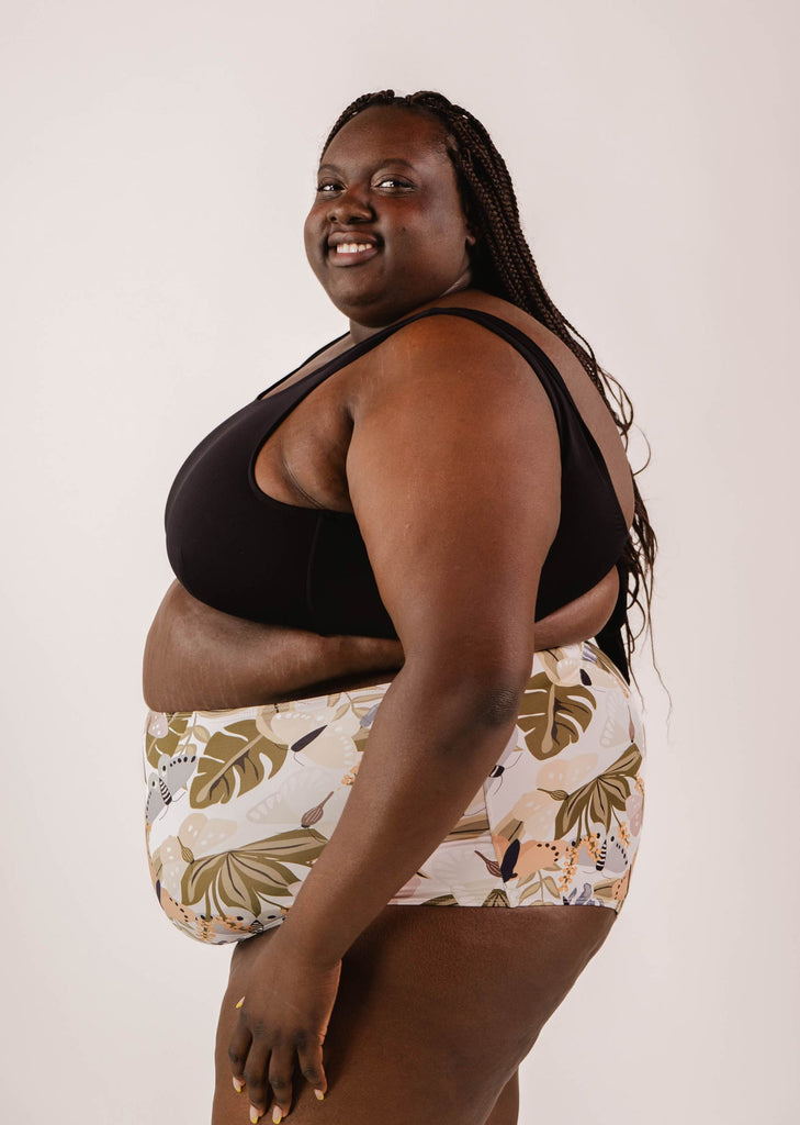 A person smiling and posing in a Mimi & August Lima Black Bralette Bikini Top with adjustable back and patterned shorts against a white background.