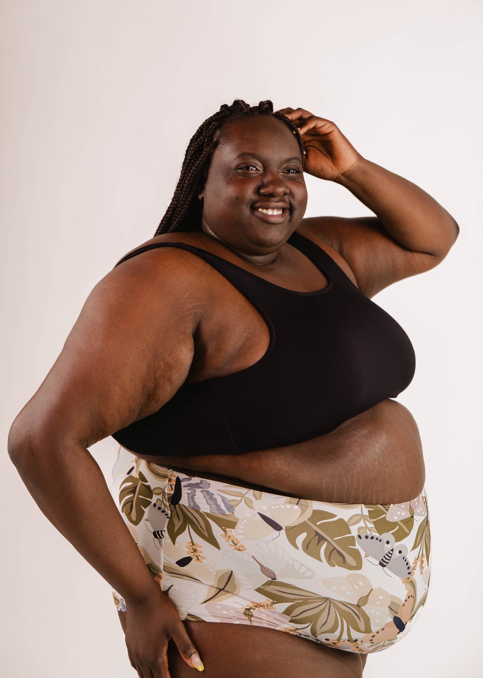 A person with braided hair smiles while posing in a Lima Black Bralette Bikini Top by Mimi & August, featuring adjustable back and removable pads, paired with patterned shorts against a plain background.