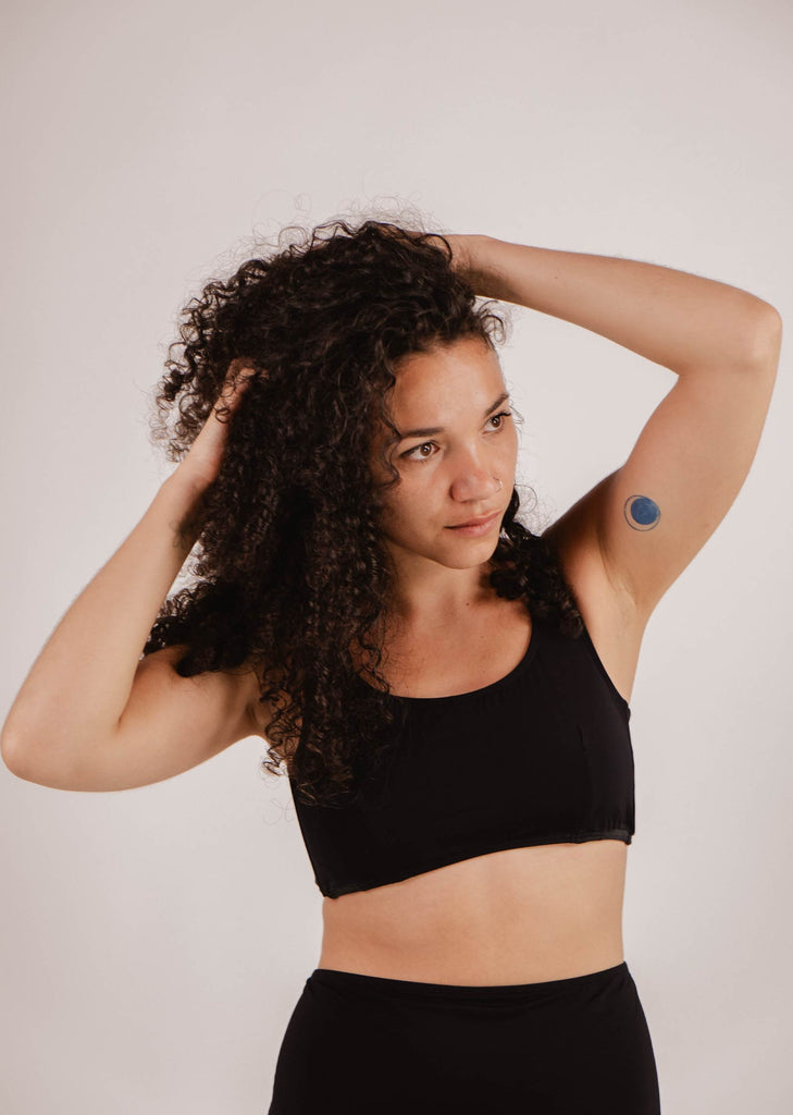 A person with curly hair wearing a Mimi & August Lima Black Bralette Bikini Top and black bottoms touches their hair. There is a small blue circular patch on the upper part of their left arm.