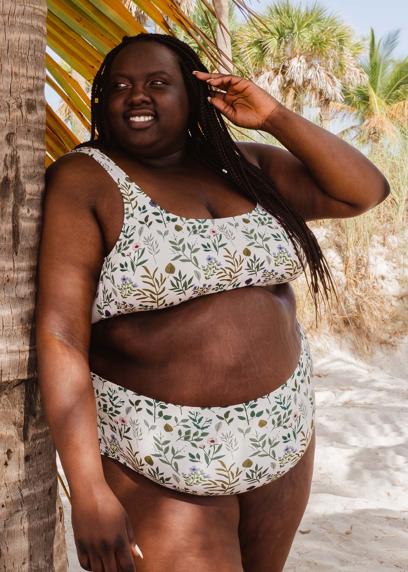 Femme africaine vêtue d'un haut de bikini mimi et august Lima Herboria Bralette, debout à côté de palmiers.