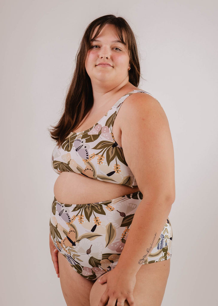 A person with long hair is wearing a patterned two-piece swimsuit, featuring the Lima Mariposa Bralette Bikini Top by Mimi & August with an adjustable back, and standing against a plain background, radiating comfort and confidence.