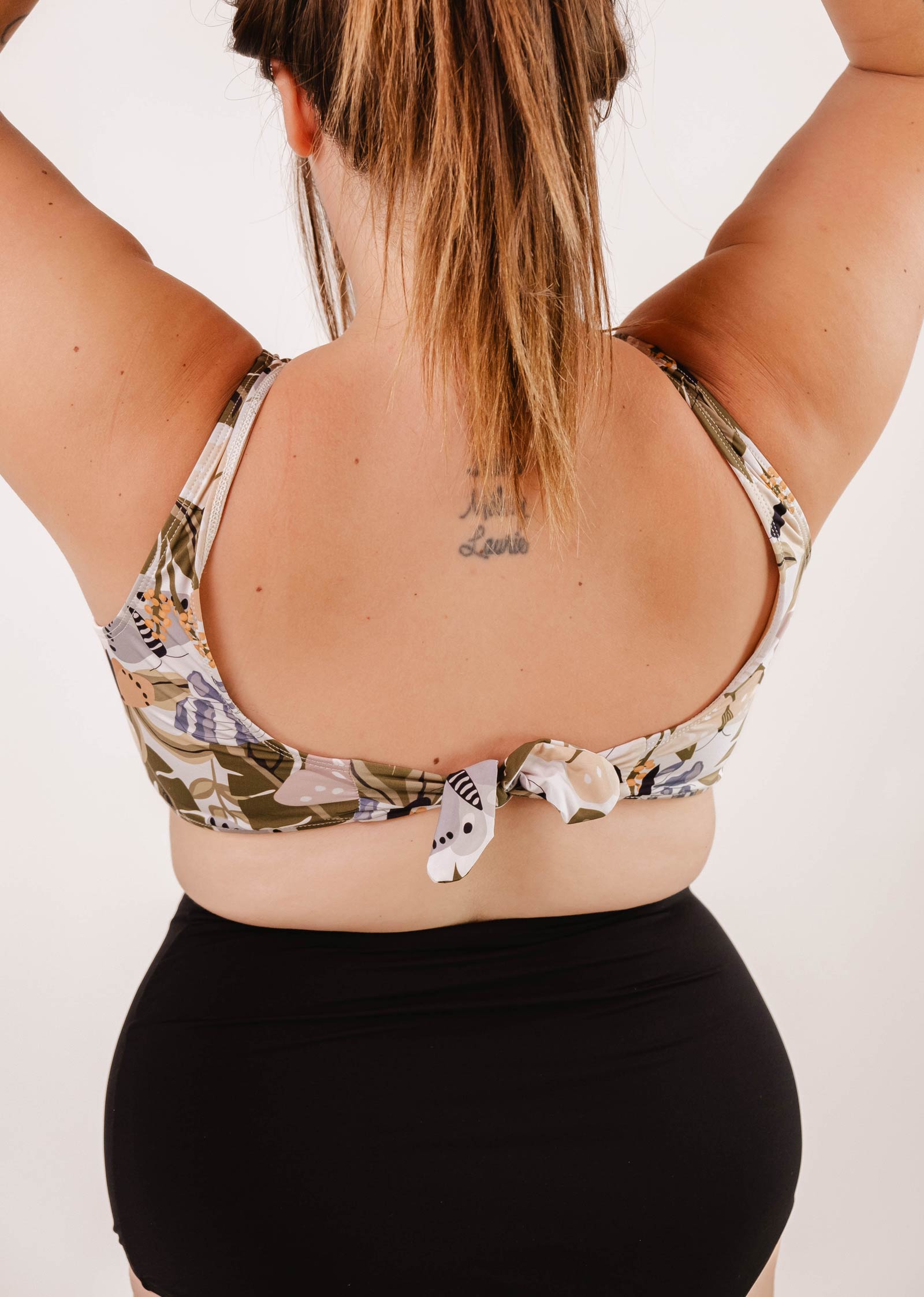 La personne ayant un tatouage dans le dos porte un haut de bikini Mimi & August Lima Mariposa Bralette à motifs floraux et un bas de bikini noir, debout, les bras levés pour plus de confort et de confiance.