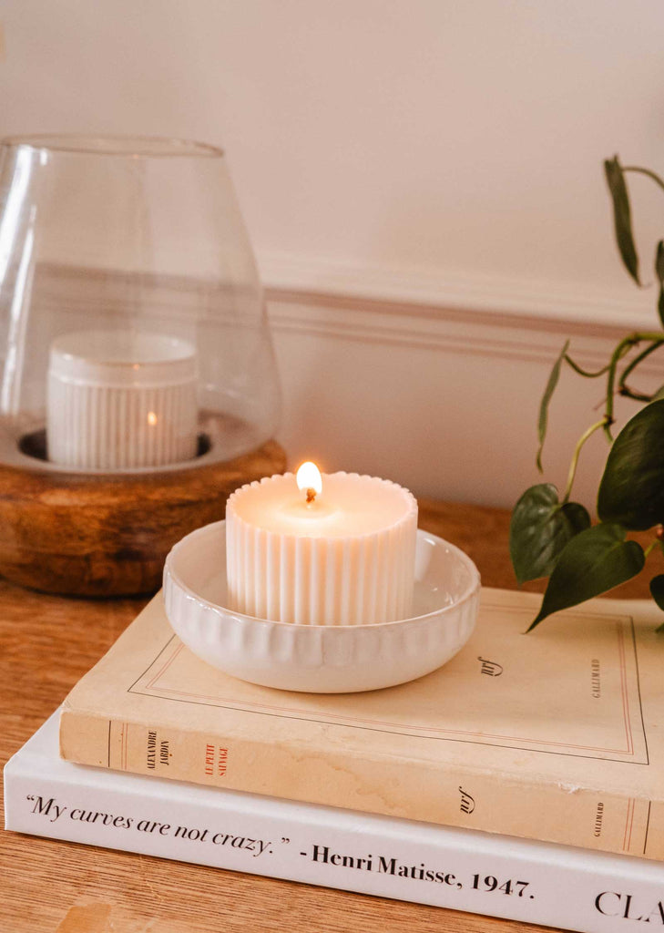 A lit Little Ceramic Nest by Mimi & August sits on a pile of books, one of which reads "My curves are not crazy. - Henri Matisse, 1947." A plant is visible in the background.