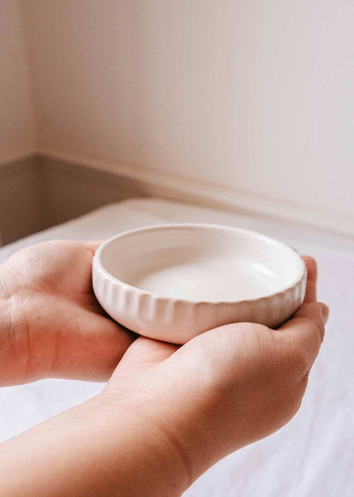 A pair of hands holding a small, white, round handcrafted ceramic plate, known as the Little Ceramic Nest by Mimi & August, against a light background.