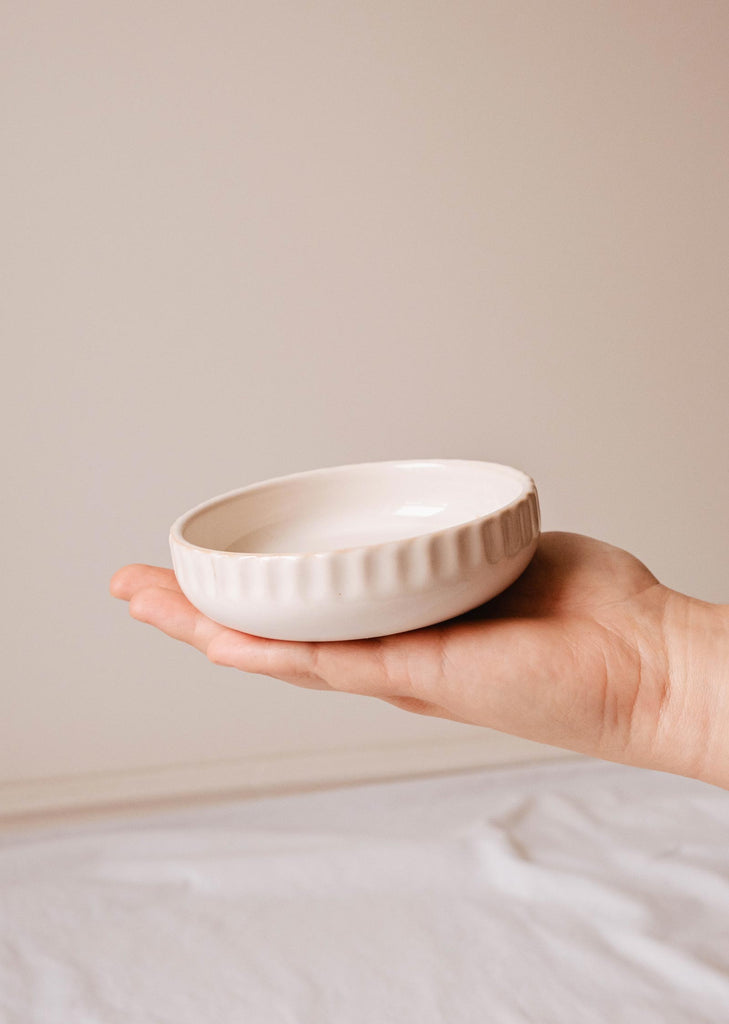 Person holding a Little Ceramic Nest, a small, white ceramic bowl with a ribbed exterior edge handcrafted by Mimi & August, over a white surface.