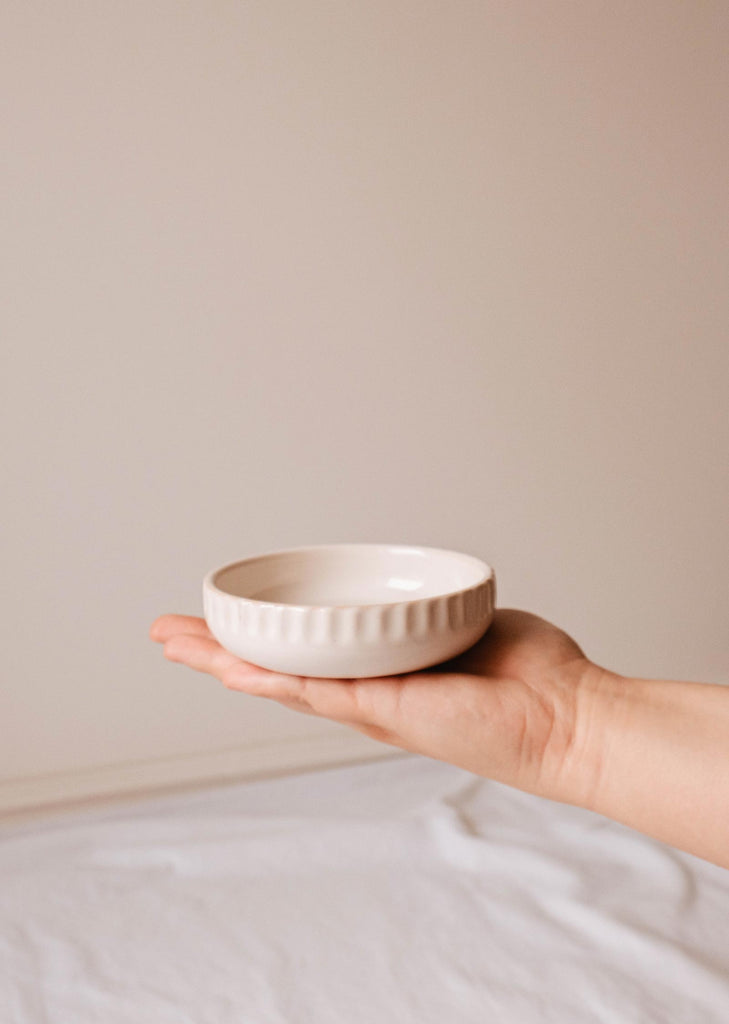 A hand holding a small, white, round ceramic dish with a ridged exterior against a neutral background. The handcrafted Little Ceramic Nest by Mimi & August suggests it could easily double as an elegant candle holder.