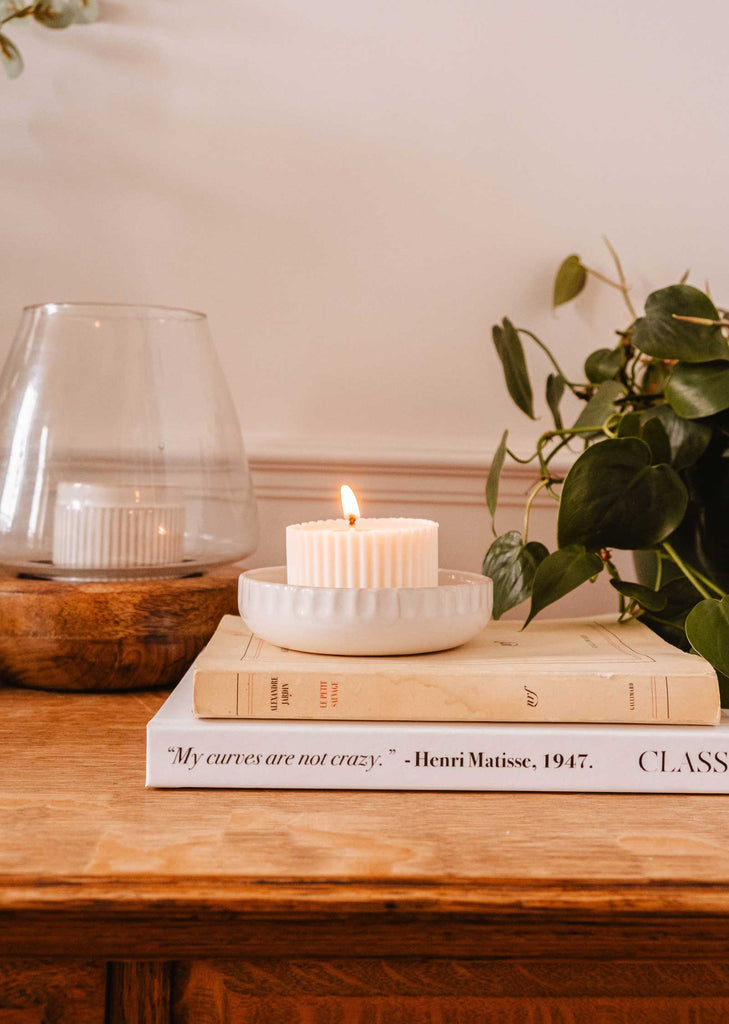 A lit candle in the Little Ceramic Nest by Mimi & August is placed on top of two stacked books beside a plant and a large glass container on a wooden surface.