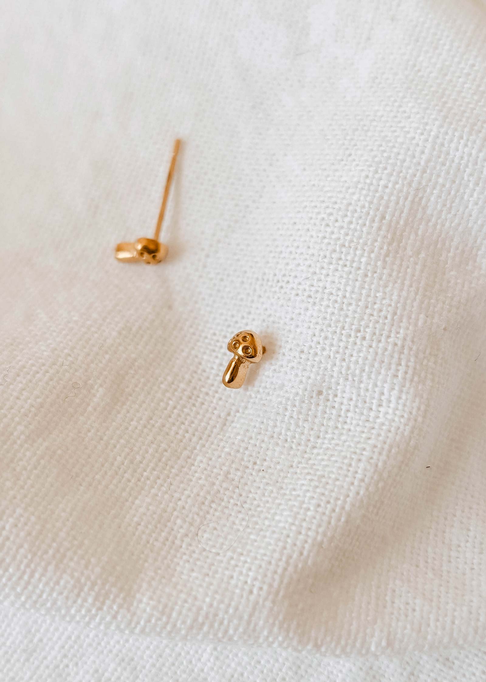 These charming gold-plated earrings, crafted by Mimi & August and named "Little Mushroom," elegantly rest on a white fabric surface, showcasing their unique handmade-in-USA quality.