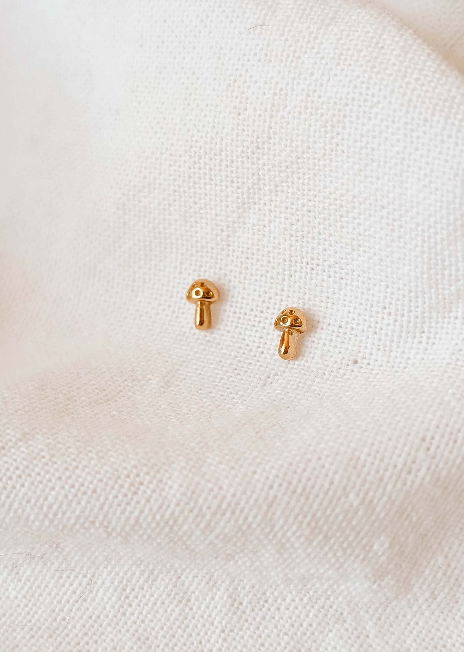 Les boucles d'oreilles en plaqué or Little Mushroom de Mimi & August, fabriquées à la main aux États-Unis, scintillent délicatement sur un fond de tissu blanc texturé.