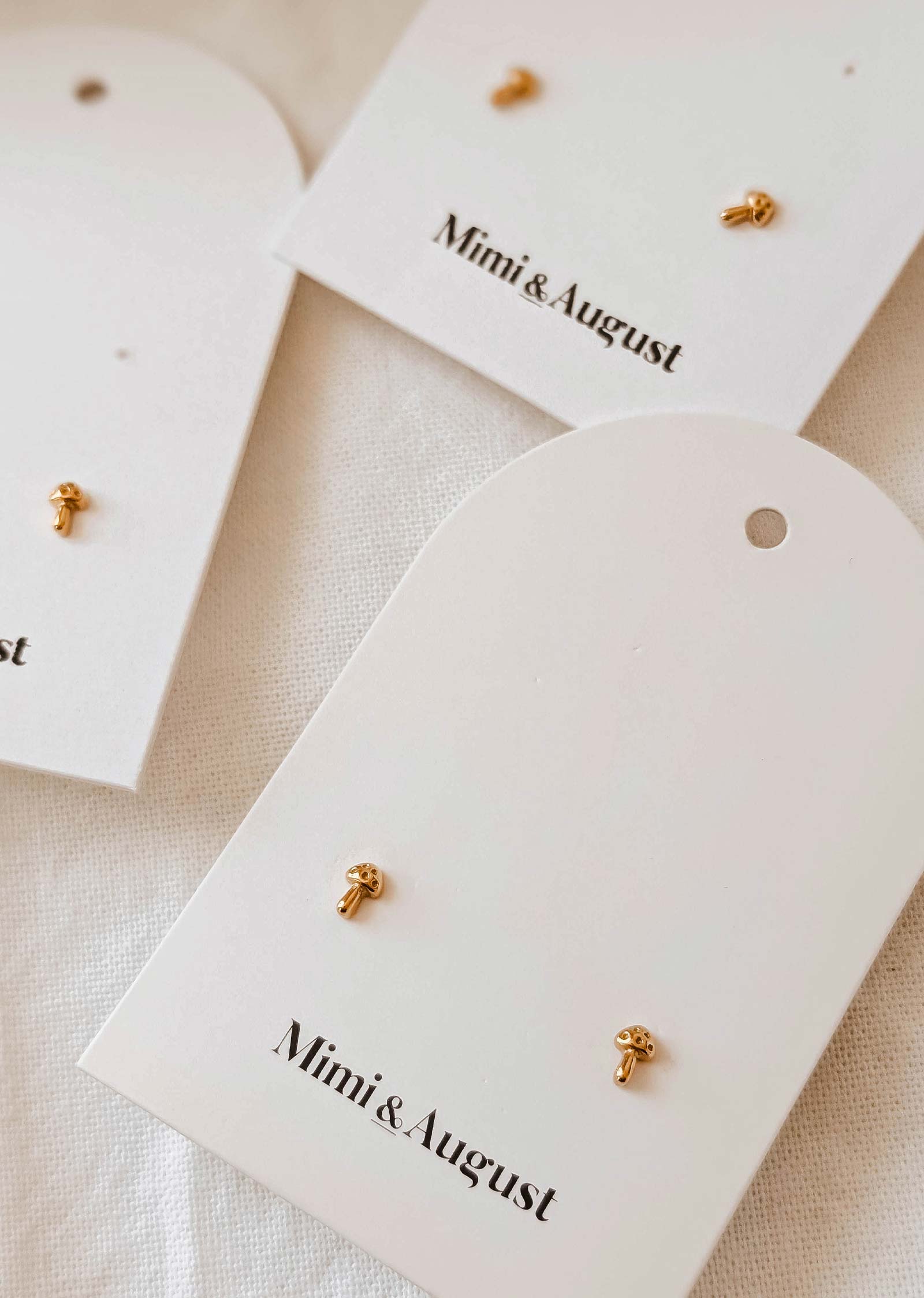 Three pairs of gold-plated Little Mushroom earrings by Mimi & August are elegantly displayed on white cards.