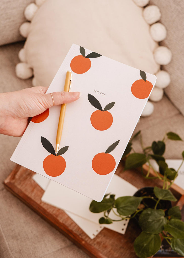 A person holding a Mimi & August notebook decorated with Little Oranges for note-taking.