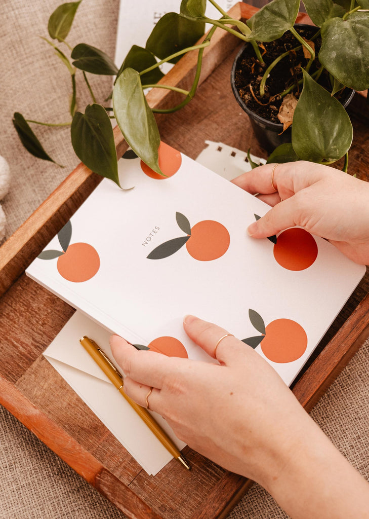 A hand holding a Mimi & August notebook adorned with Little Oranges for note-taking.