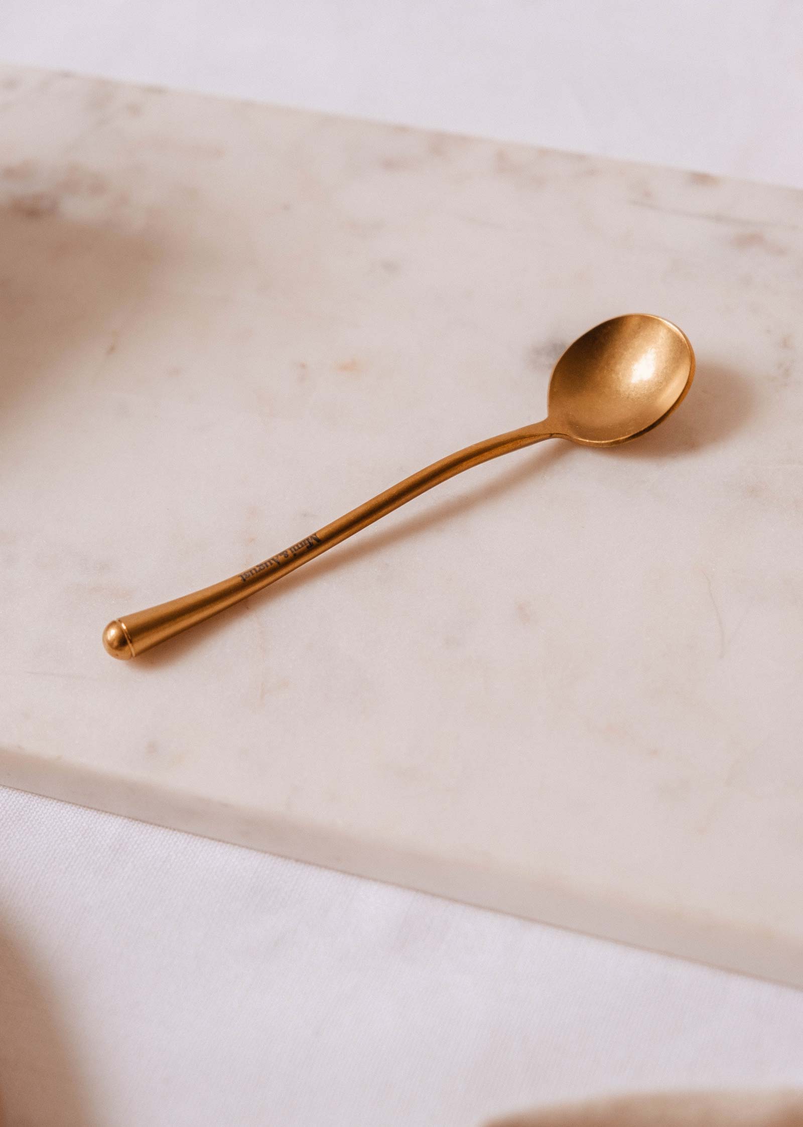 The Golden Touch – Delicate Gold Teaspoon by Mimi & August shines atop a white marble surface.