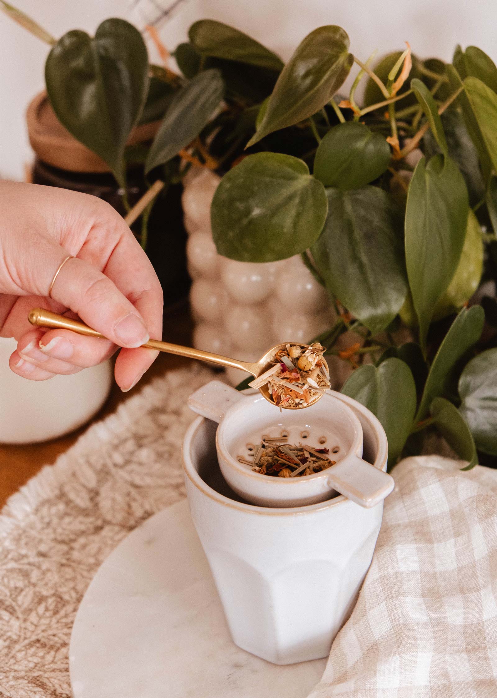 Une main tient The Golden Touch - Delicate Gold Teaspoon de Mimi & August au-dessus d'une tasse blanche avec une passoire à thé, entourée de plantes vertes et d'un tissu beige.