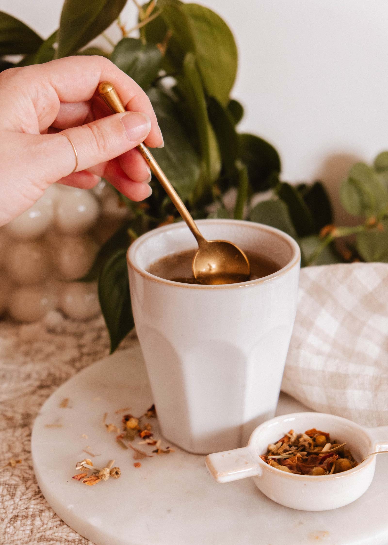 A hand stirs a warm drink in a white mug with Mimi & August's The Golden Touch – Delicate Gold Teaspoon, while loose tea leaves and a small white dish sit on marble, framed by lush green plants.