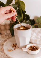 Une main remue une boisson chaude dans une tasse blanche avec cette cuillère à thé délicate en or de Mimi & August, tandis que des feuilles de thé en vrac et un petit plat blanc sont posés sur du marbre, encadrés par des plantes vertes luxuriantes.