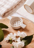 Le Nid d'Infusion - passoire à thé en céramique de Mimi & August repose sur une surface en bois entourée de fleurs blanches et de feuilles vertes, avec un tissu plié et d'autres céramiques en arrière-plan, créant ainsi une scène paisible d'infusion de thé.