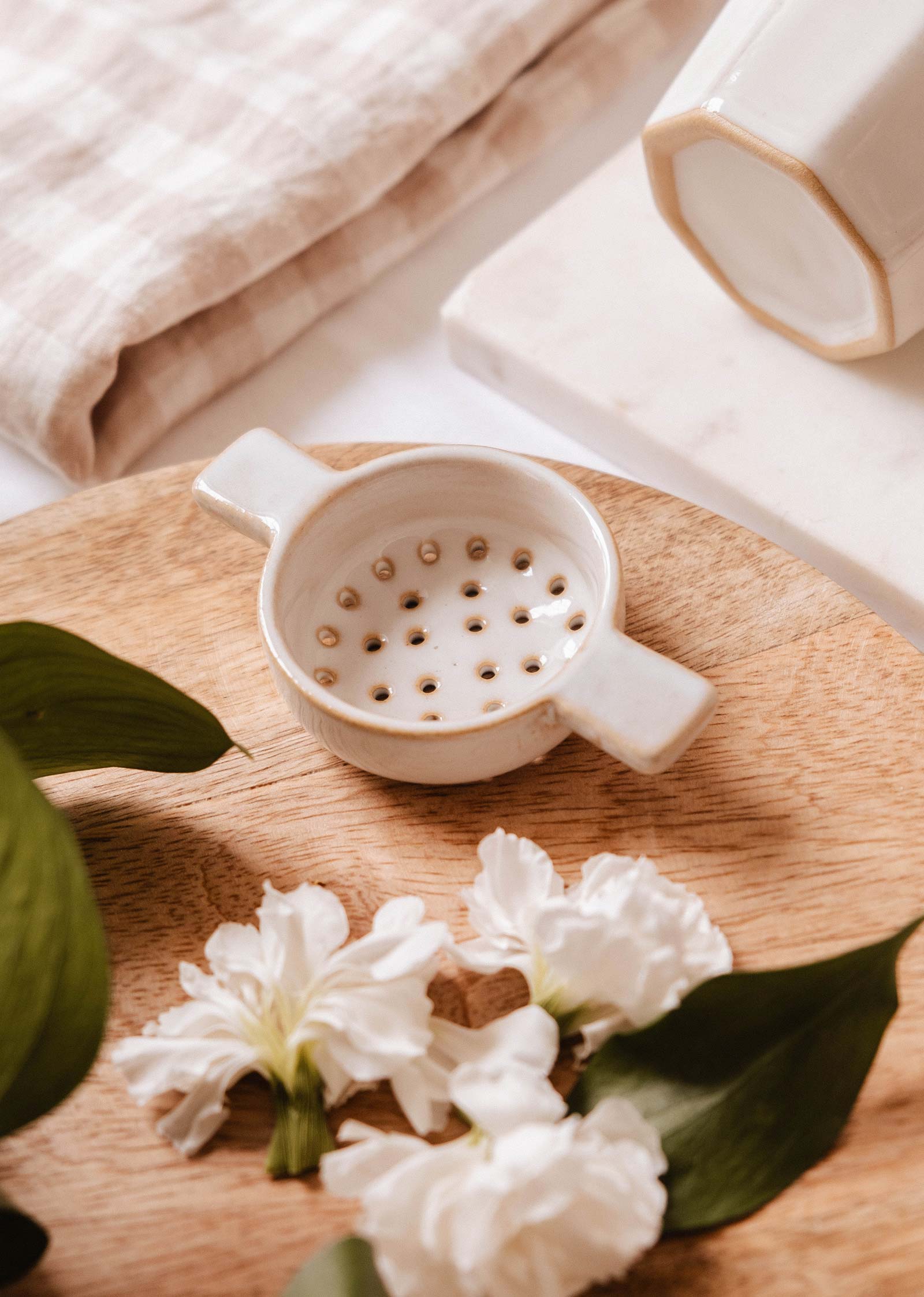 Le Nid d'Infusion - passoire à thé en céramique de Mimi & August repose sur une surface en bois entourée de fleurs blanches et de feuilles vertes, avec un tissu plié et d'autres céramiques en arrière-plan, créant ainsi une scène paisible d'infusion de thé.