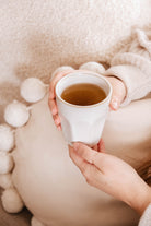 Une personne vêtue d'un pull léger tient la Douceur du matin tasse en ceramique de Mimi & August, remplie de thé, tout en étant assise contre un coussin texturé avec des accents de pompon. La tasse fabriquée à la main passe au micro-ondes et au lave-vaisselle.
