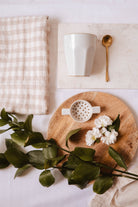 Le coffret Little Moments de Mimi & August comprend une tasse blanche, une cuillère dorée et une passoire à thé en céramique sur marbre, avec un torchon en lin et de délicates fleurs blanches à feuilles vertes disposées sur une planche en bois.