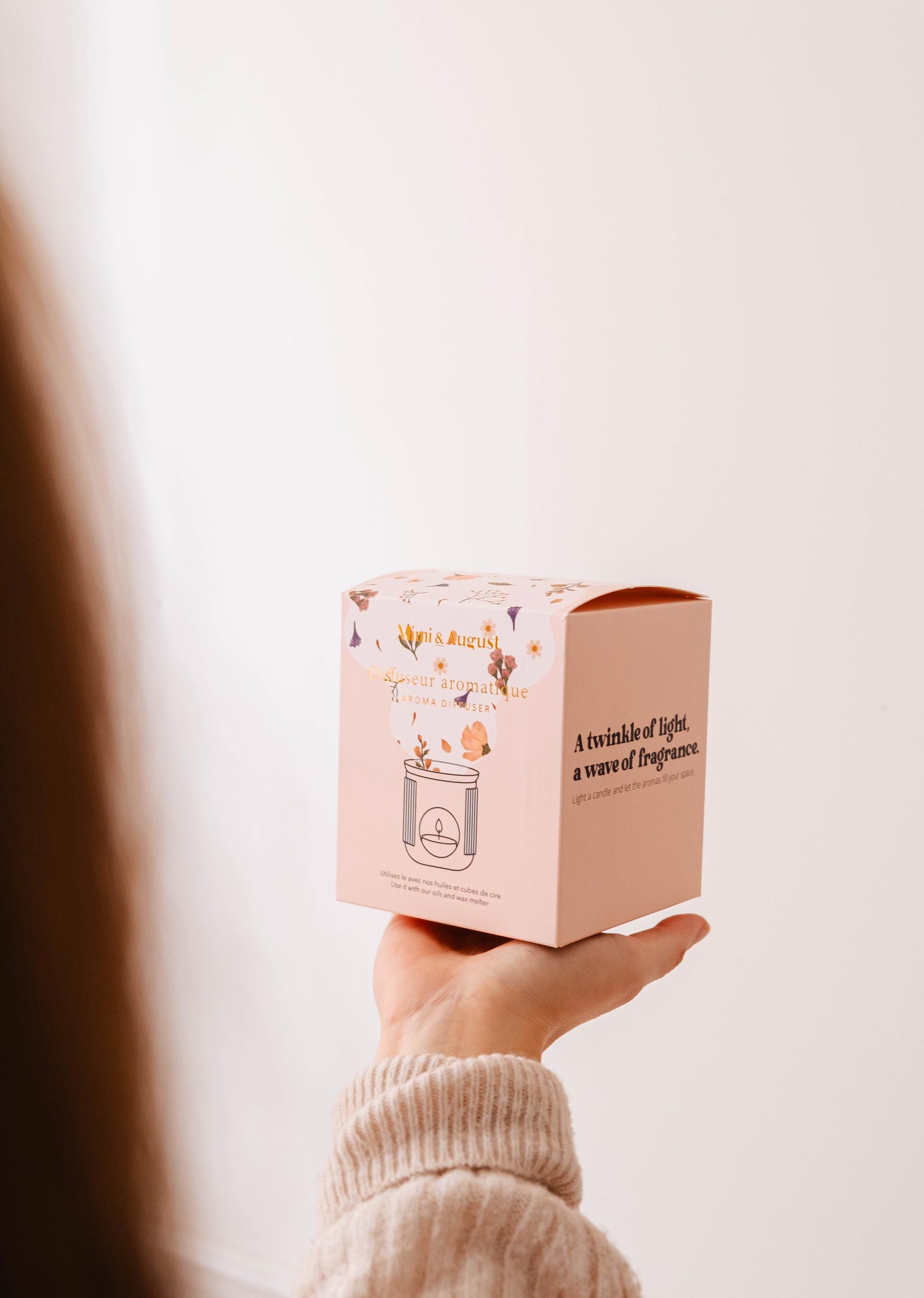 A person holds a decorative box featuring floral designs and fragrance text, along with the Lueur – Aromatic Ceramic Diffuser by Mimi & August, set against a neutral background.