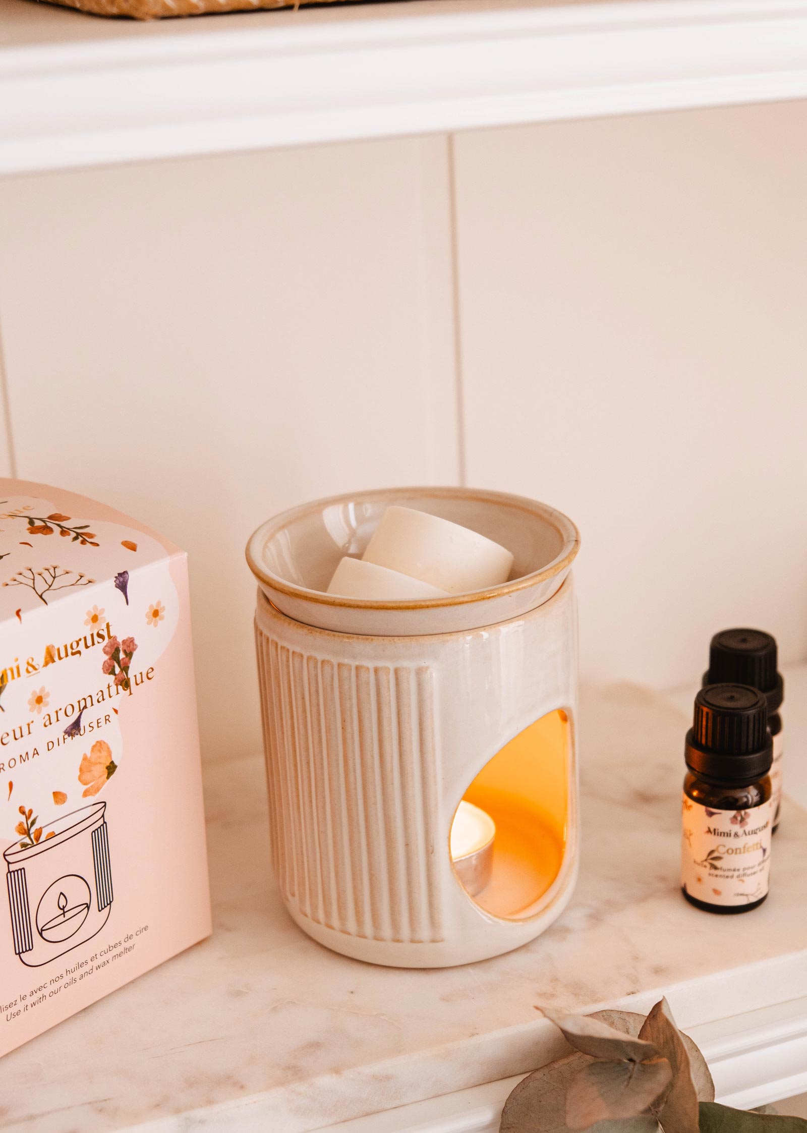 The Lueur Aromatic Ceramic Diffuser by Mimi & August, featuring a lit candle inside, is surrounded by wax melts, scented oils, and a decorative box on a shelf.