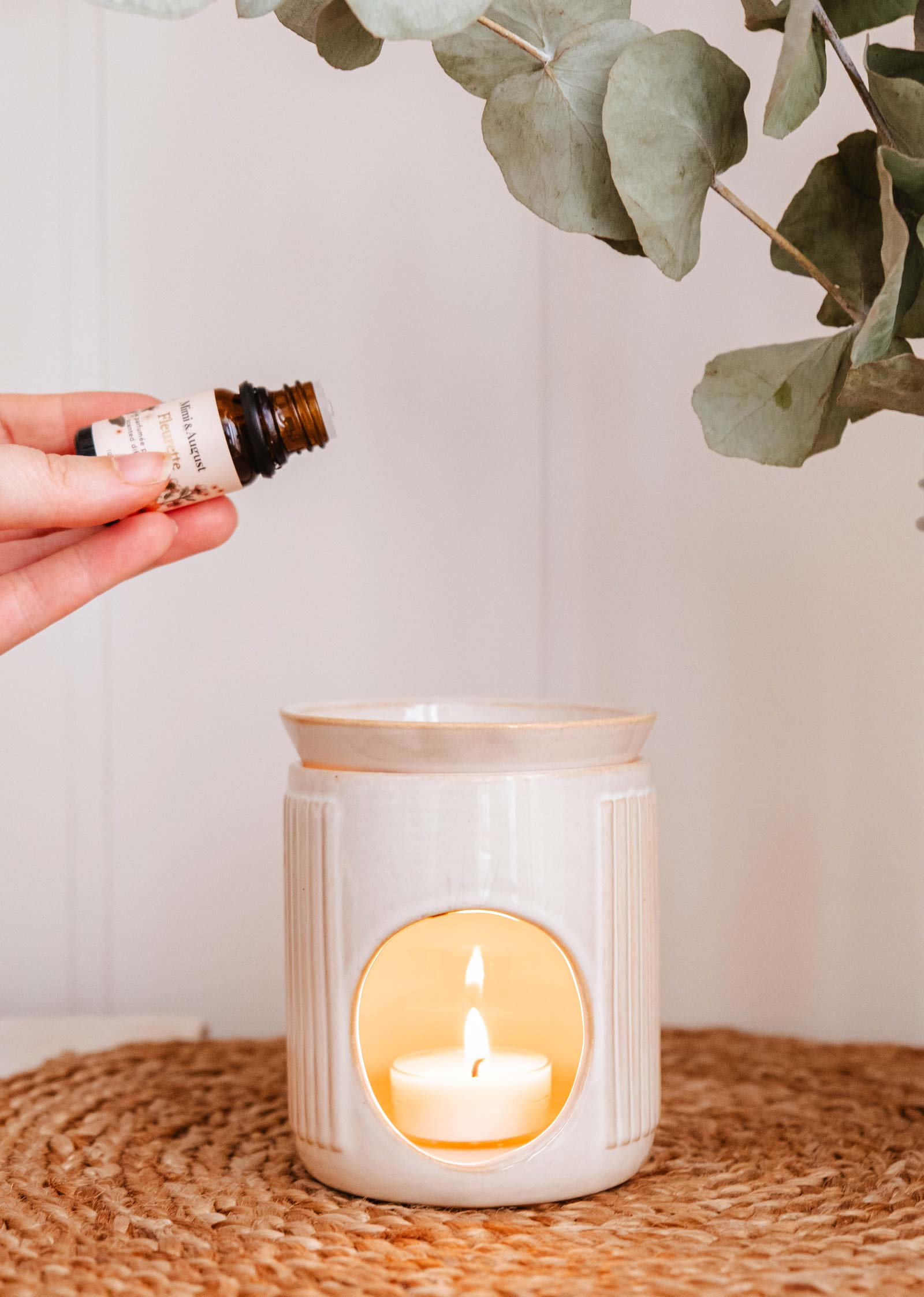 A hand holds an essential oil bottle above a lit Lueur – Aromatic Ceramic Diffuser by Mimi & August on a woven mat, surrounded by eucalyptus leaves.