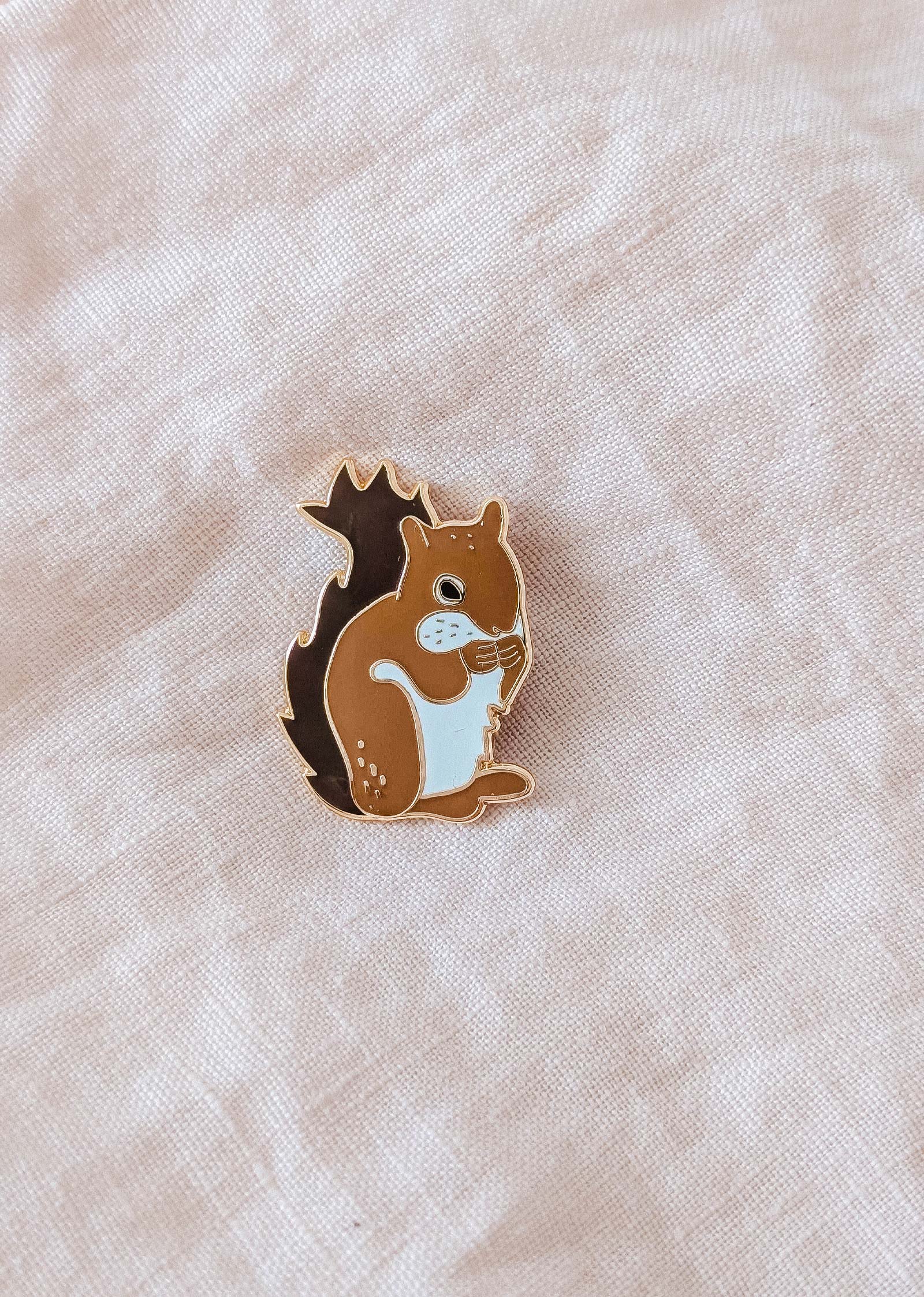 The Mimi & August MTL Squirrel Enamel Pin, in brown and white, rests on a light beige fabric—an adorable Montreal-inspired accessory ideal for animal lovers.