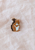 The Mimi & August MTL Squirrel Enamel Pin, in brown and white, rests on a light beige fabric—an adorable Montreal-inspired accessory ideal for animal lovers.