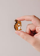 A hand displays the Mimi & August MTL Squirrel Enamel Pin—a charming brown squirrel accessory, ideal for animal lovers in Montreal—against a plain, light background.