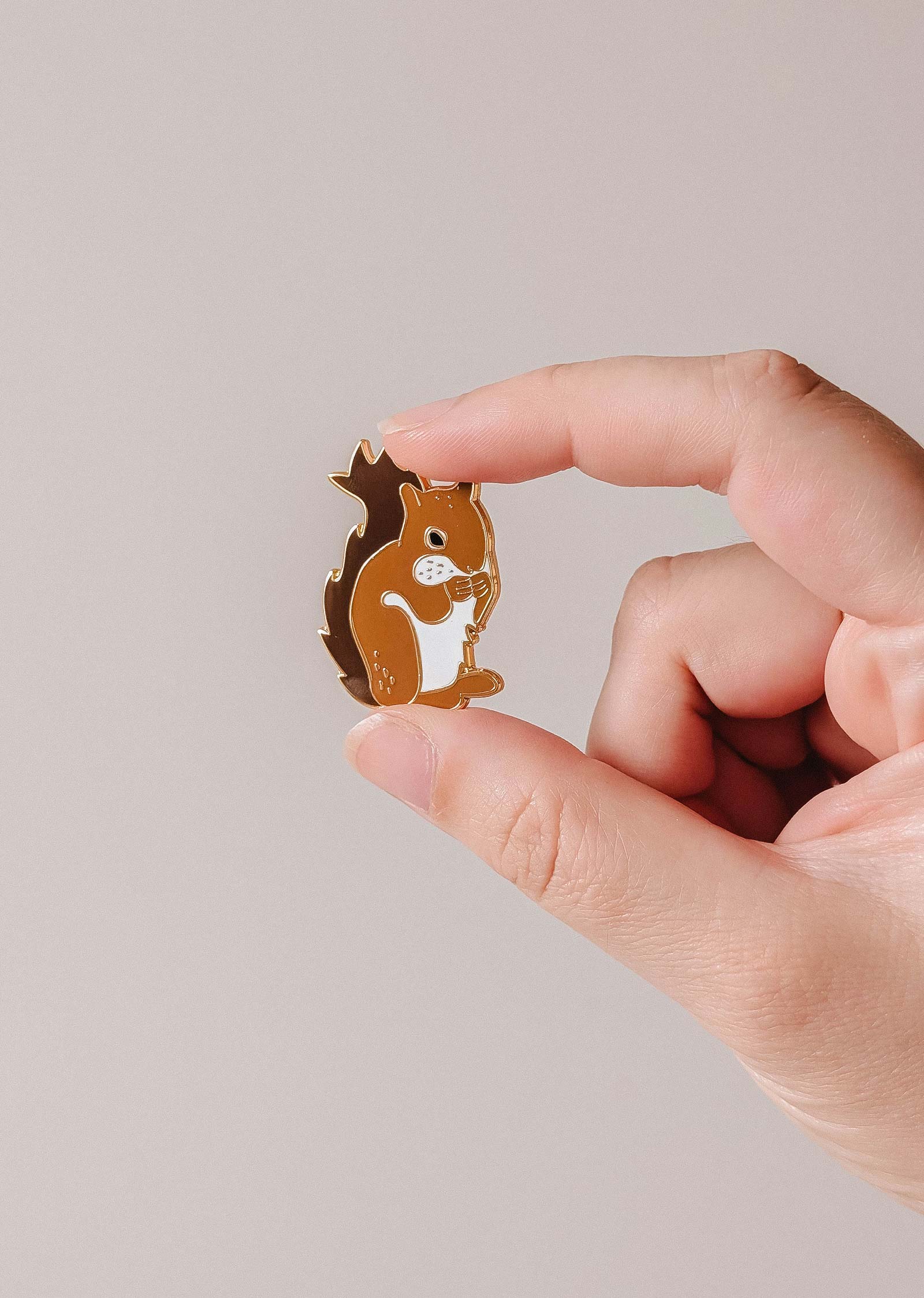 A hand displays the Mimi & August MTL Squirrel Enamel Pin—a charming brown squirrel accessory, ideal for animal lovers in Montreal—against a plain, light background.