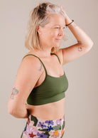 A mature woman with blonde hair in a Mimi & August Mango Amazonia Bralette Bikini Top and colorful leggings, posing with her hand on her head against a beige background.