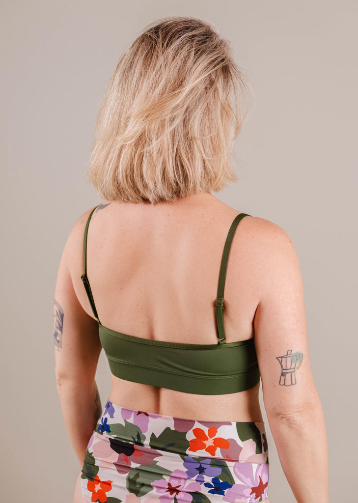 Woman with blonde bob haircut, wearing a green Mimi & August Mango Amazonia Bralette Bikini Top and floral leggings, viewed from the back. She has visible tattoos on her arms and upper back.