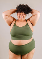 A confident woman in a Mimi & August Mango Amazonia Bralette Bikini Top and leggings, smiling as she poses with her hands lifting her curly hair.