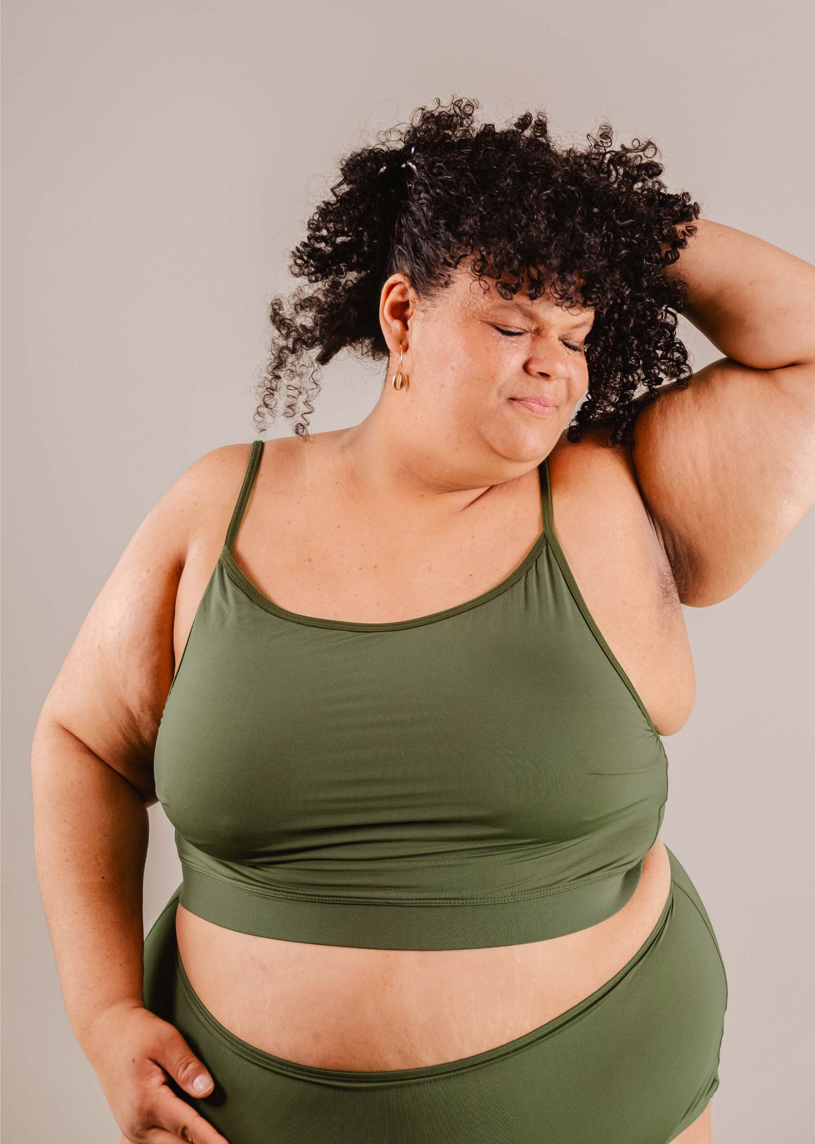 A plus-size woman in a Mimi & August Mango Amazonia Bralette Bikini Top posing with her hand on her head, her eyes closed and a slight smile, against a neutral background.