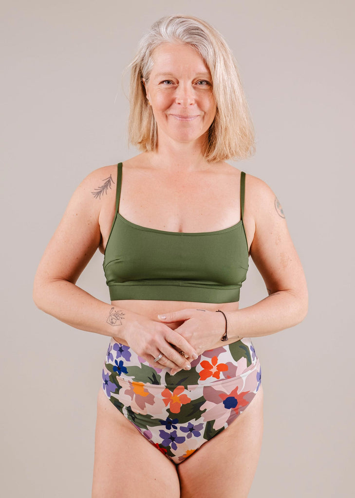 Une femme avec des cheveux gris longs sourit, vêtue d'un haut de maillot Mango Amazonia Bralette vert à bretelles réglables et d'un bas de bikini imprimé floral coloré de Mimi & August. Elle a des tatouages visibles sur les bras.