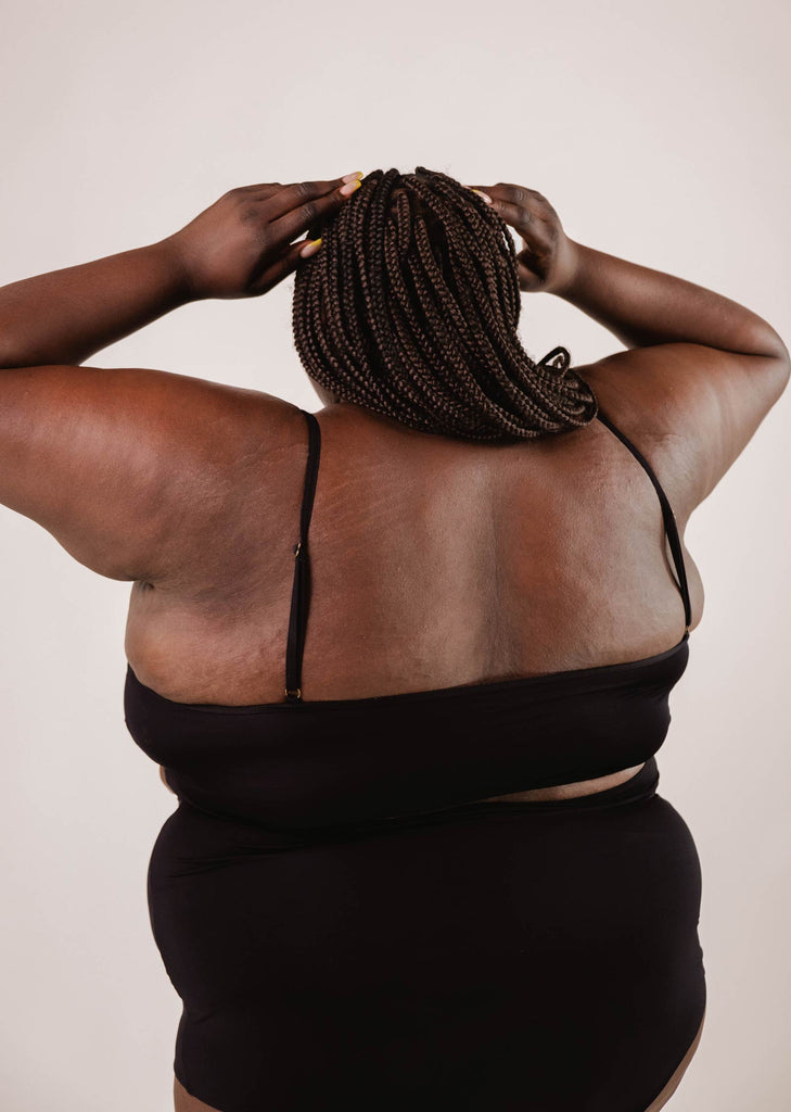 A person wearing a Mimi & August Mango Black Bralette Bikini Top with adjustable straps faces away from the camera, their hands on their head and hair styled in braids.