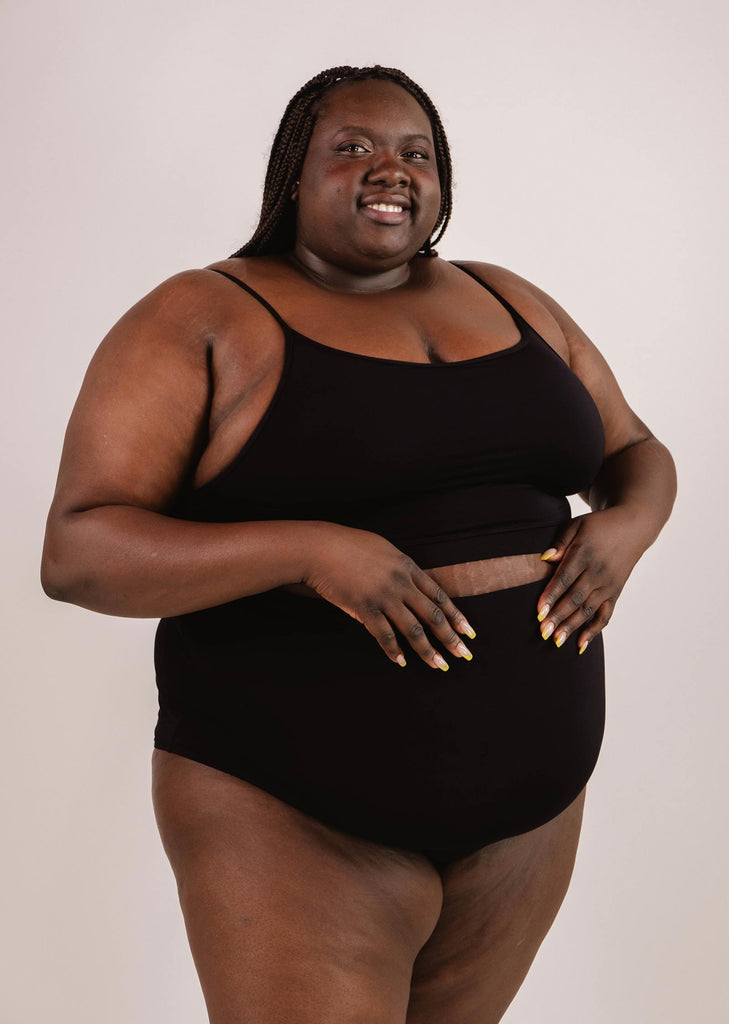 A person wearing a Mimi & August Mango Black Bralette Bikini Top and black shorts stands against a plain background, smiling and looking at the camera. They have braided hair and are holding the edge of their top with their hands, showcasing the adjustable straps that offer excellent support.