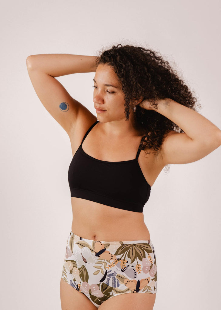 A person with curly hair wearing a Mimi & August Mango Black Bralette Bikini Top and floral-patterned high-waisted underwear, with an armband sensor on their upper arm, stands with arms raised against a plain background.