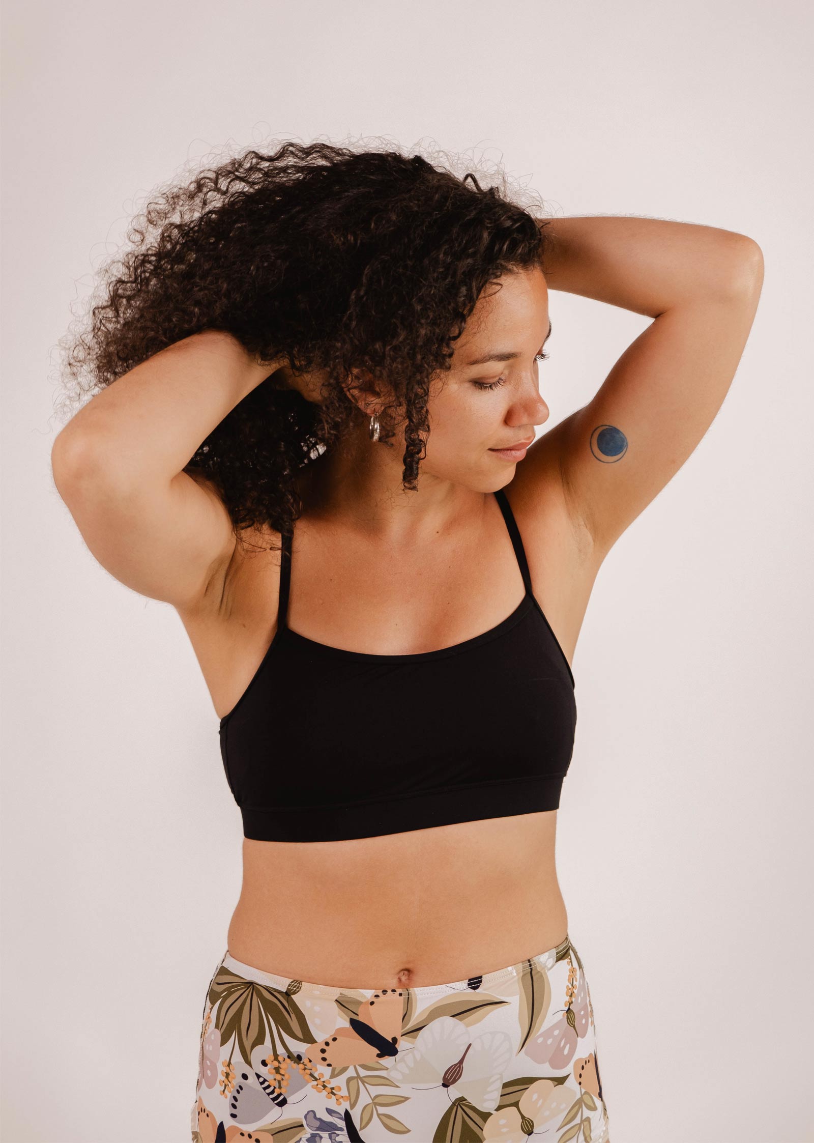 A person with curly hair, wearing a Mimi & August Mango Black Bralette Bikini Top and patterned leggings, adjusts their hair with both hands. A round adhesive patch is visible on their upper arm.